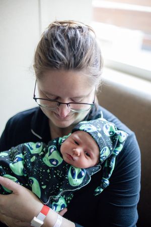 New mother with glasses lovingly gazing at her newborn in alien print swaddle and hat.