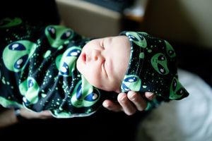 Sleeping newborn baby swaddled in green alien print blanket and matching hat held in parent's hands.