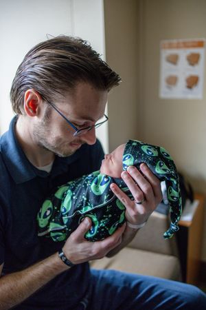 New father tenderly kissing his newborn baby dressed in alien print outfit at hospital.
