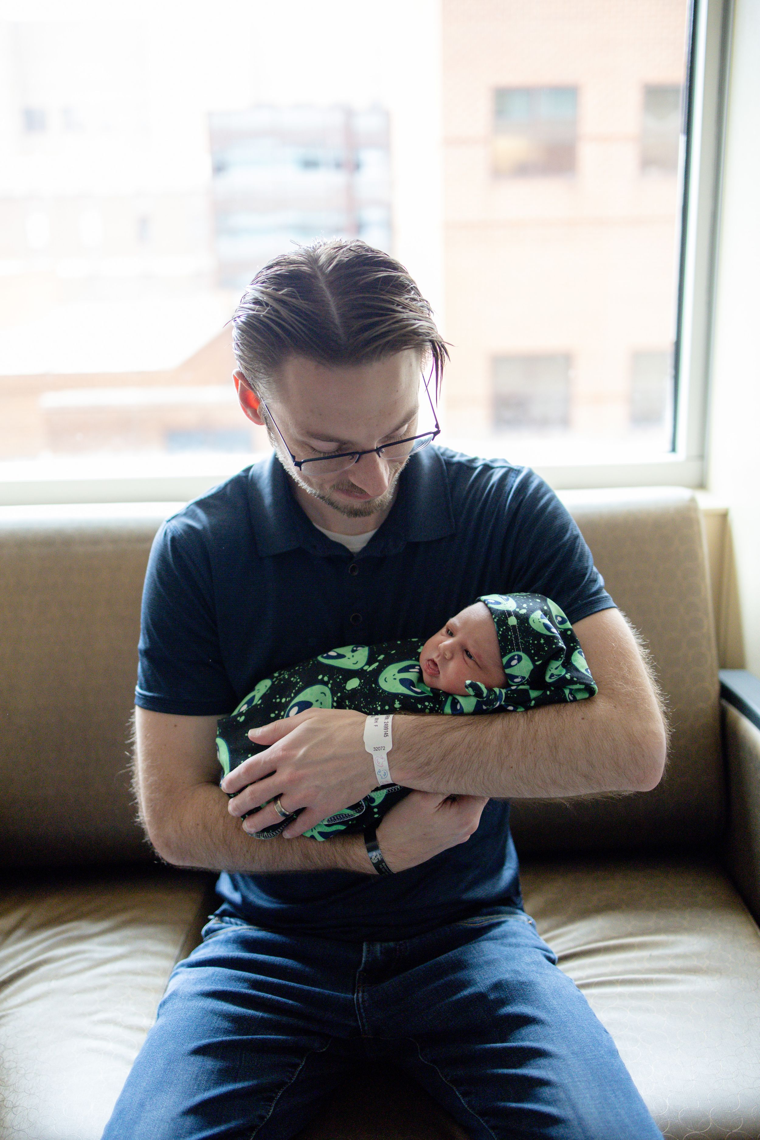 Father in navy polo shirt holding sleeping newborn baby wrapped in alien print blanket while sitting on couch.