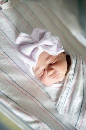 Sleeping newborn baby wrapped in striped hospital blanket wearing a white bow hat.
