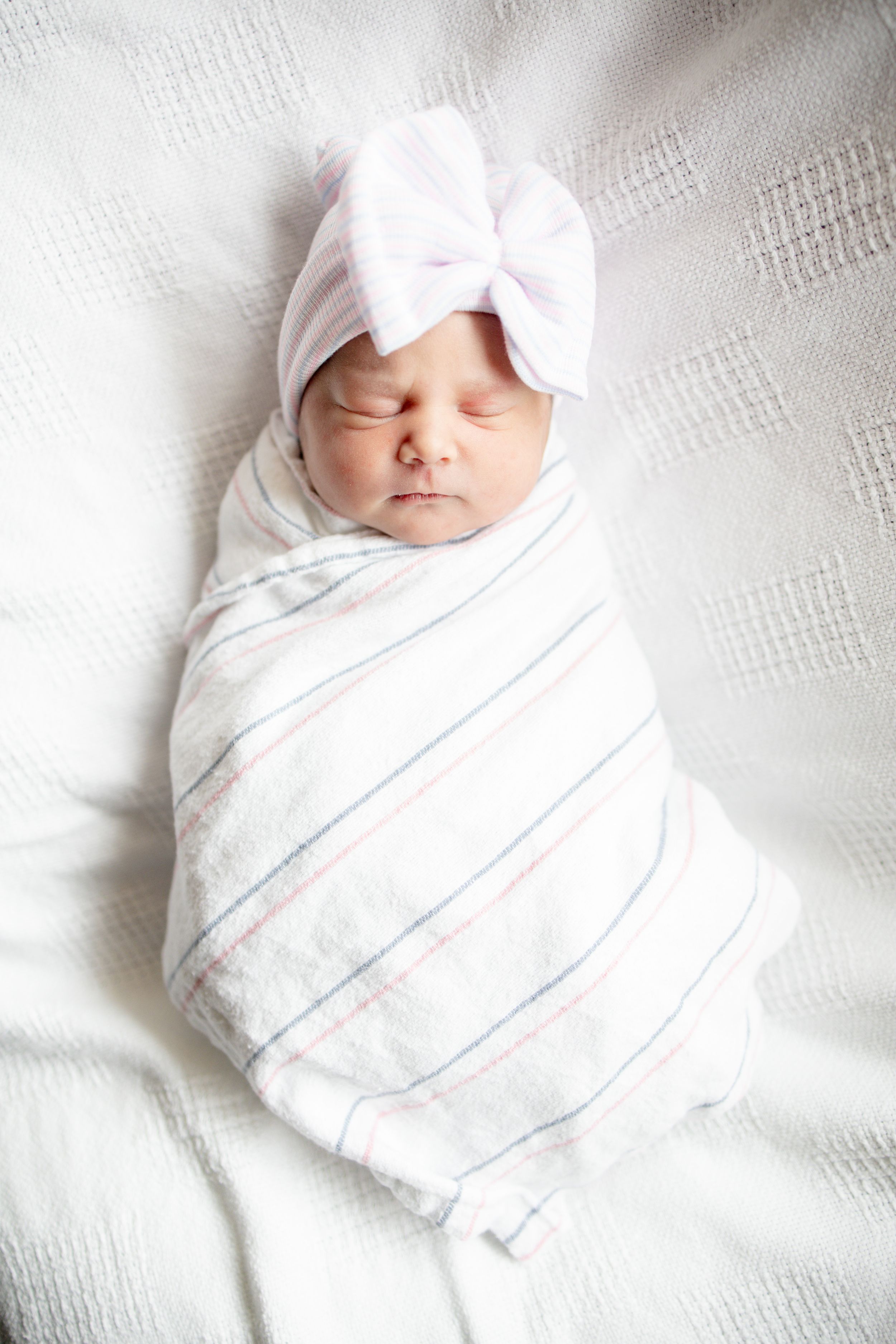 Sleeping newborn baby swaddled in striped blanket wearing white bow hat on white background.
