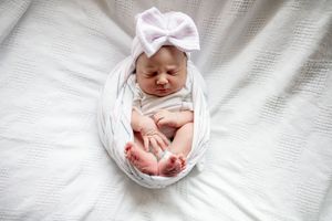 Newborn baby girl swaddled in white wrap with tiny feet peeking out wearing lavender bow headband.