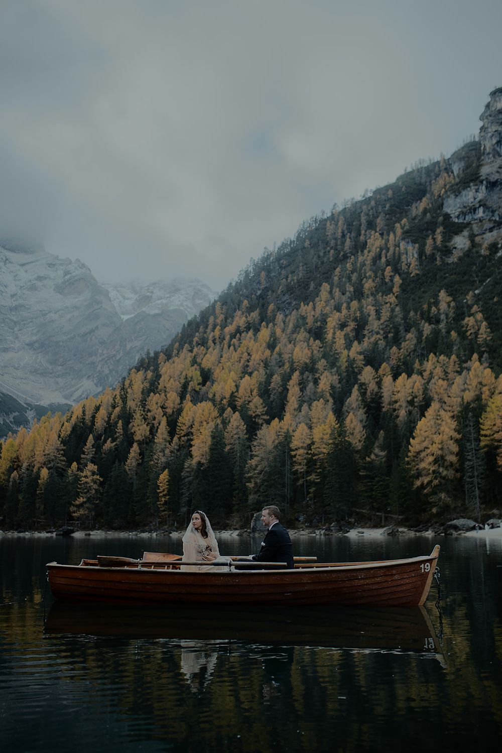 Bride and groom sitting in a red wooden boat on a calm lake surrounded by autumn trees and snowy peaks.