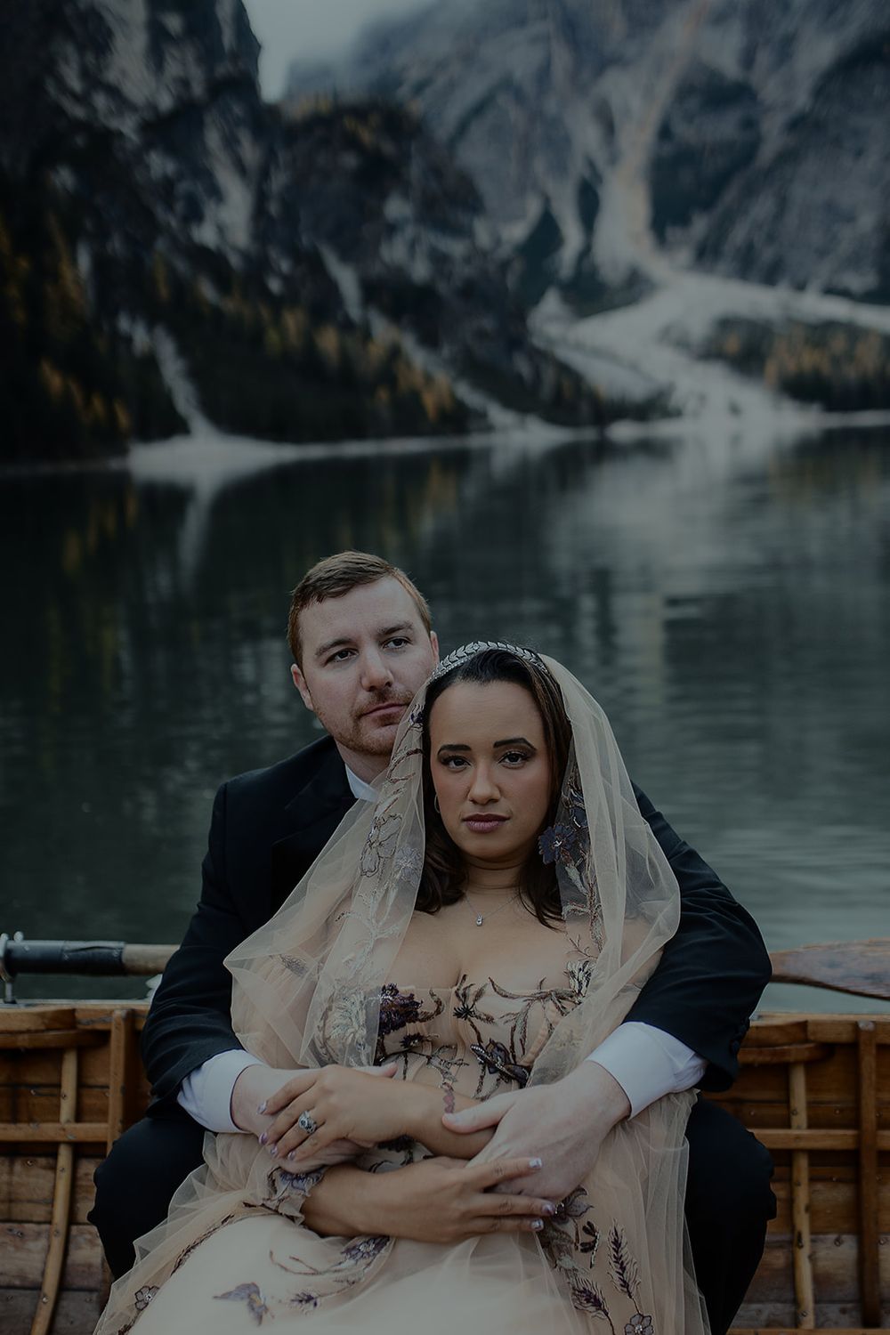 Couple embracing in a wooden boat on a serene alpine lake with misty mountains reflected in the water.