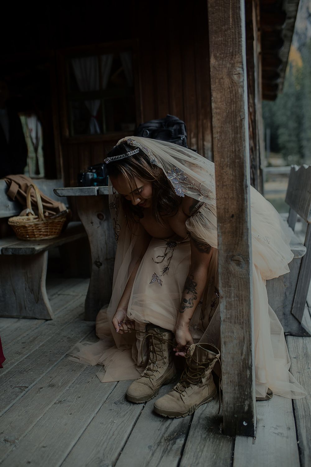 Bride in wedding dress and veil lacing up combat boots on rustic wooden porch of log cabin.