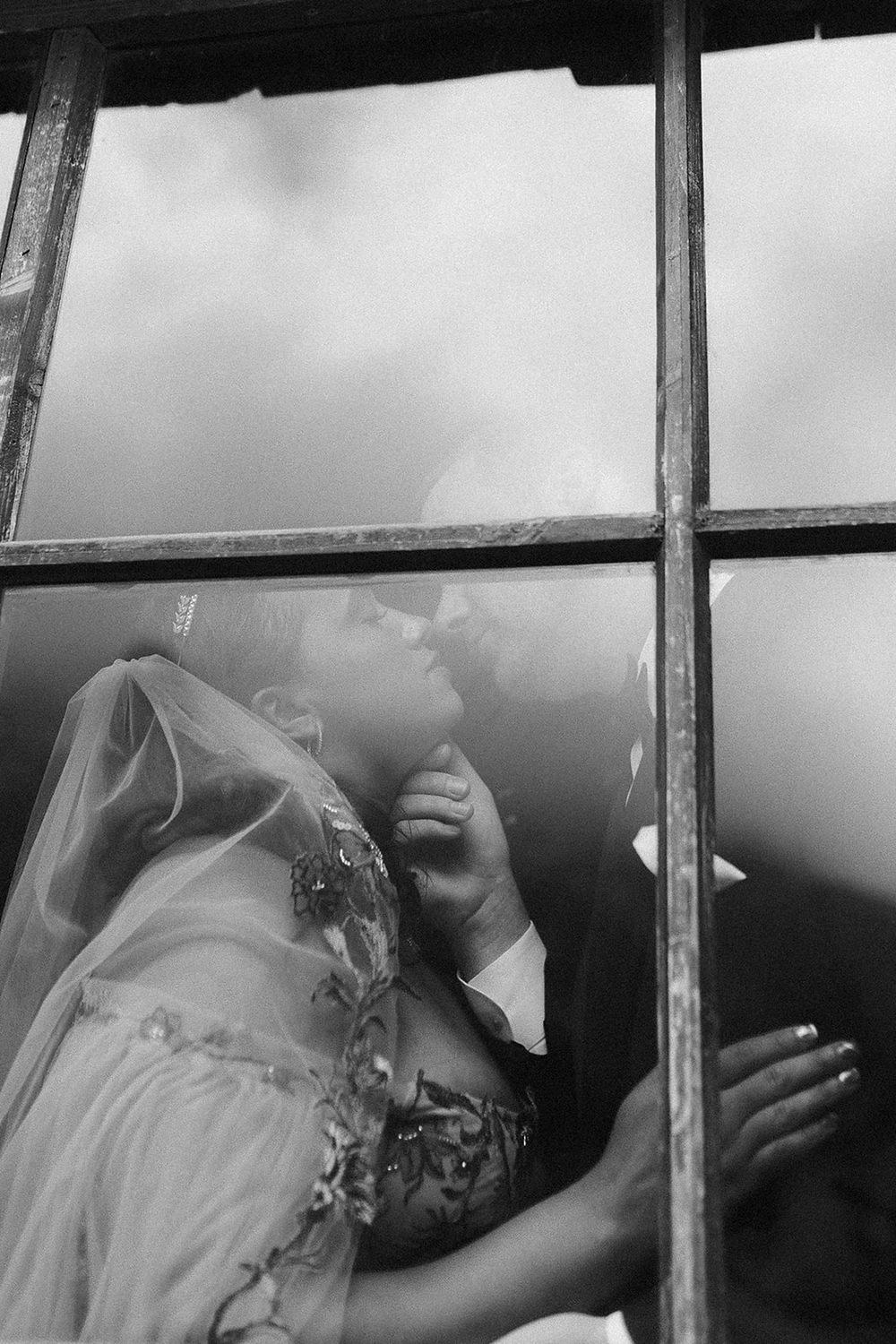 Black and white wedding photo of bride and groom kissing through a window pane.