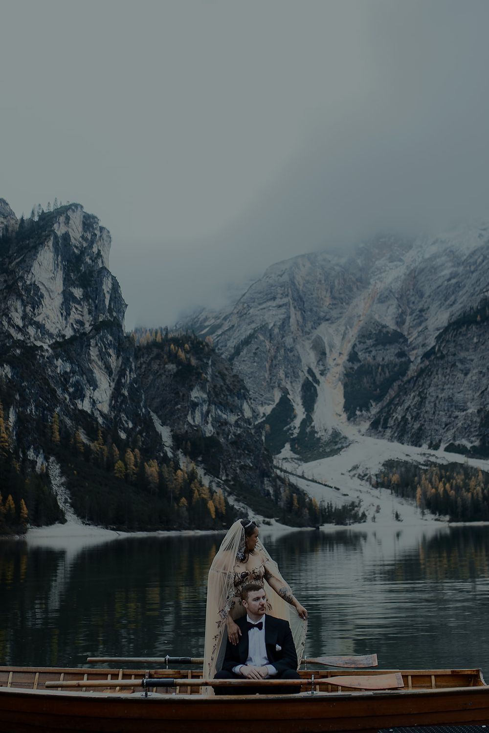 Bride and groom in a wooden boat on a serene alpine lake with misty mountains in the Dolomites.