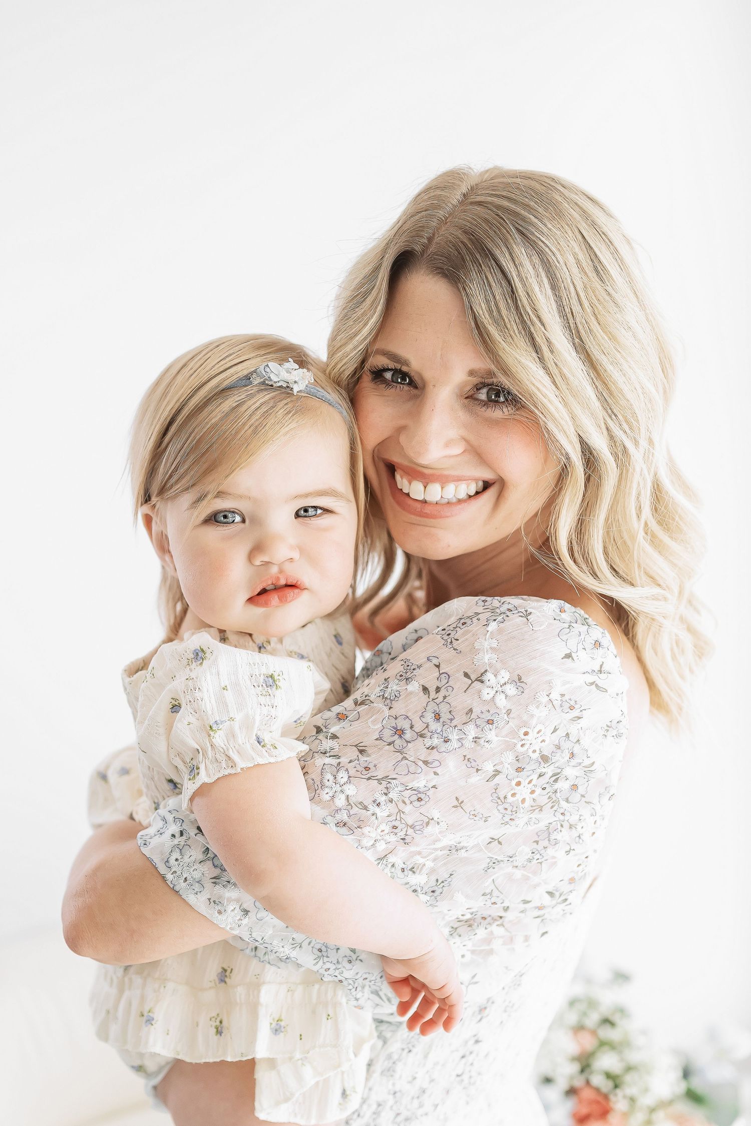 Smiling blonde woman holding a baby girl in matching white floral dresses against a white background.