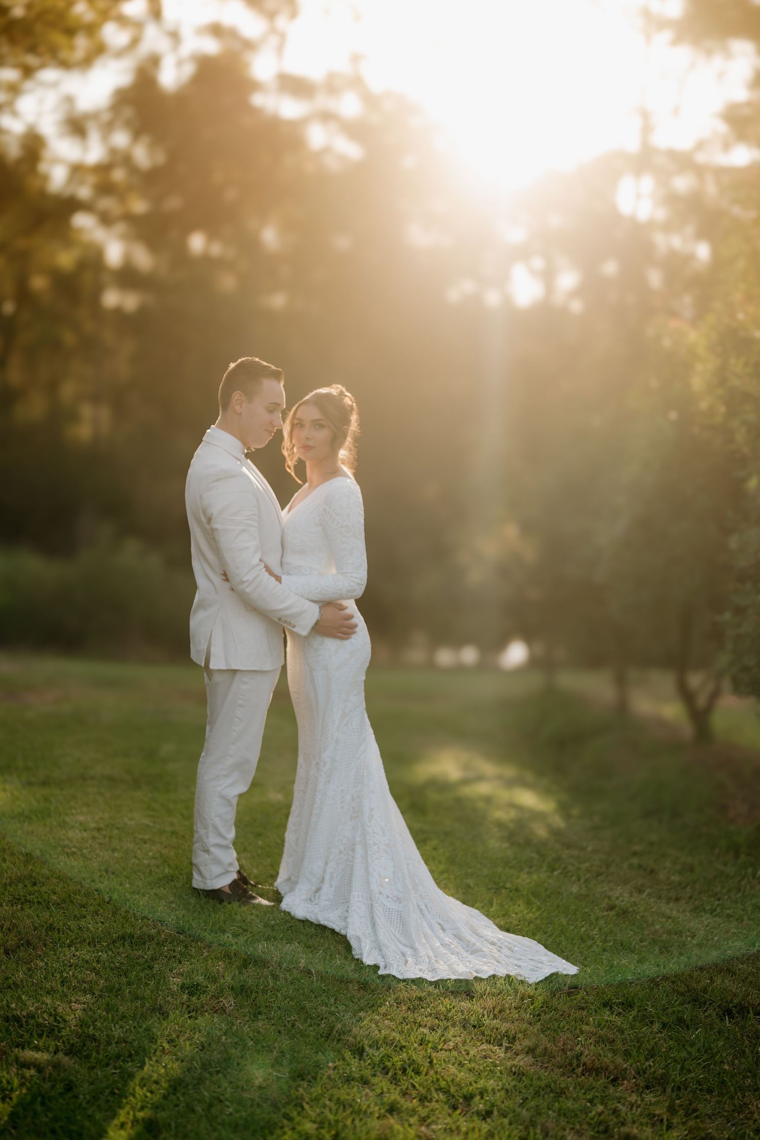 A bride and groom embrace in golden sunset light on green grass, surrounded by trees, wearing elegant white wedding attire.