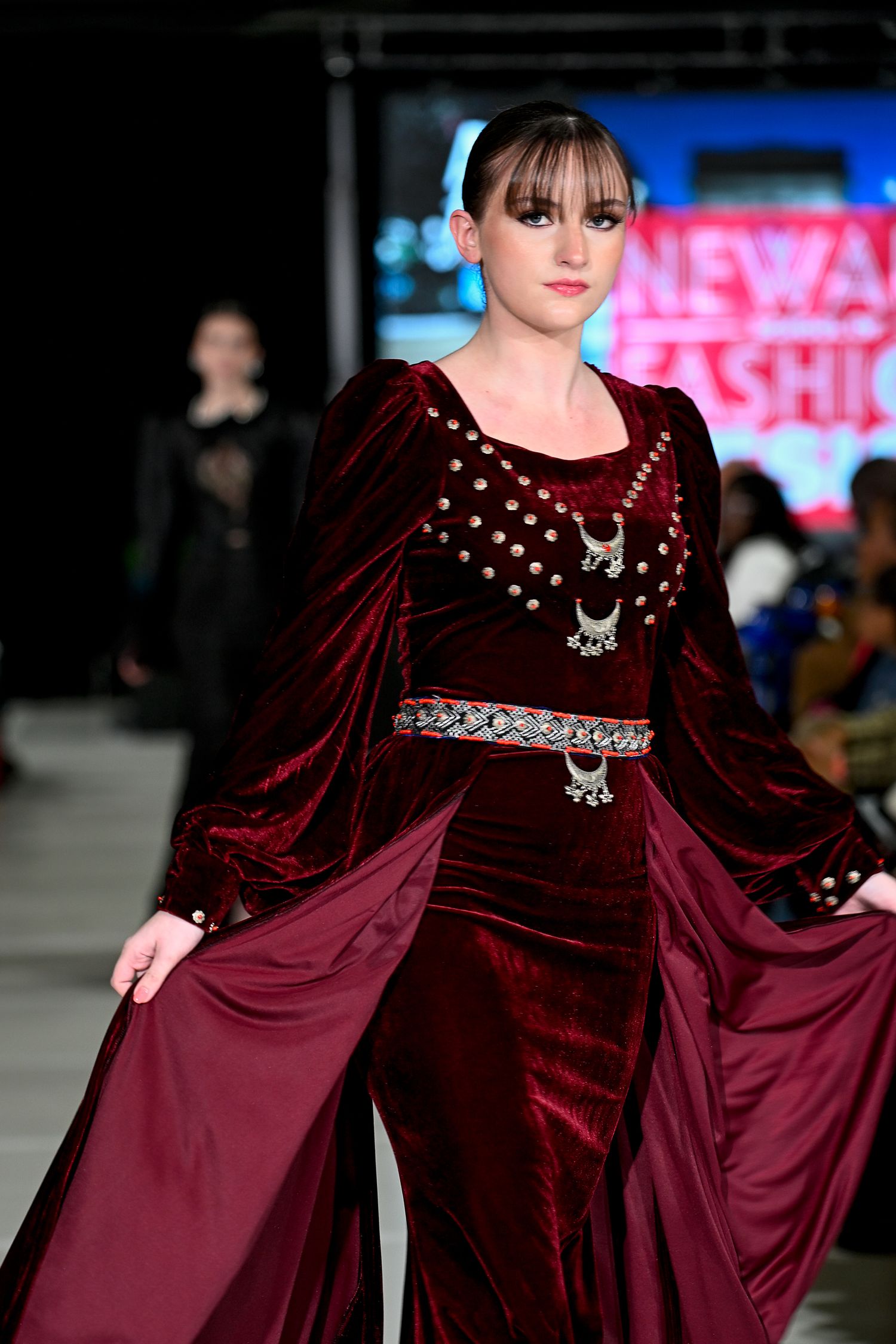 Model walks runway in burgundy velvet gown with silver embellishments and wide sleeves at a fashion show.