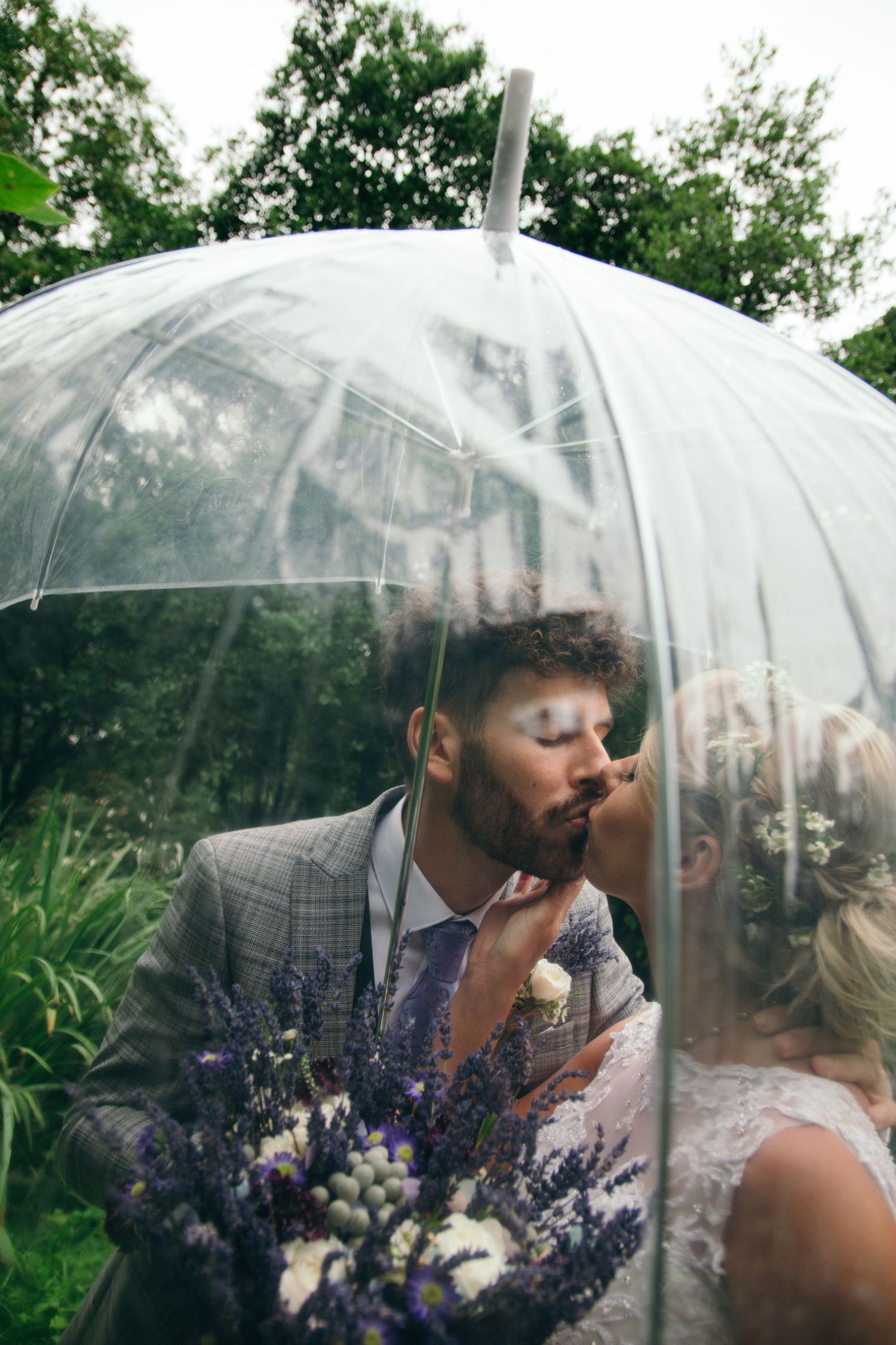Bride and groom kissing under a clear bubble umbrella, holding a lavender bouquet in a lush garden setting.