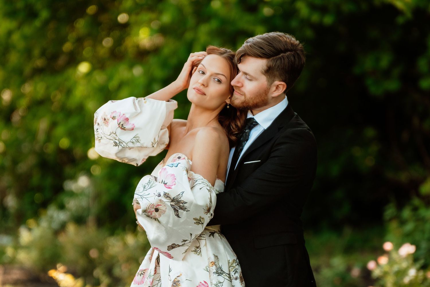 Couple embracing in garden, bride in floral off-shoulder gown, groom in black tuxedo, lush green trees backdrop.