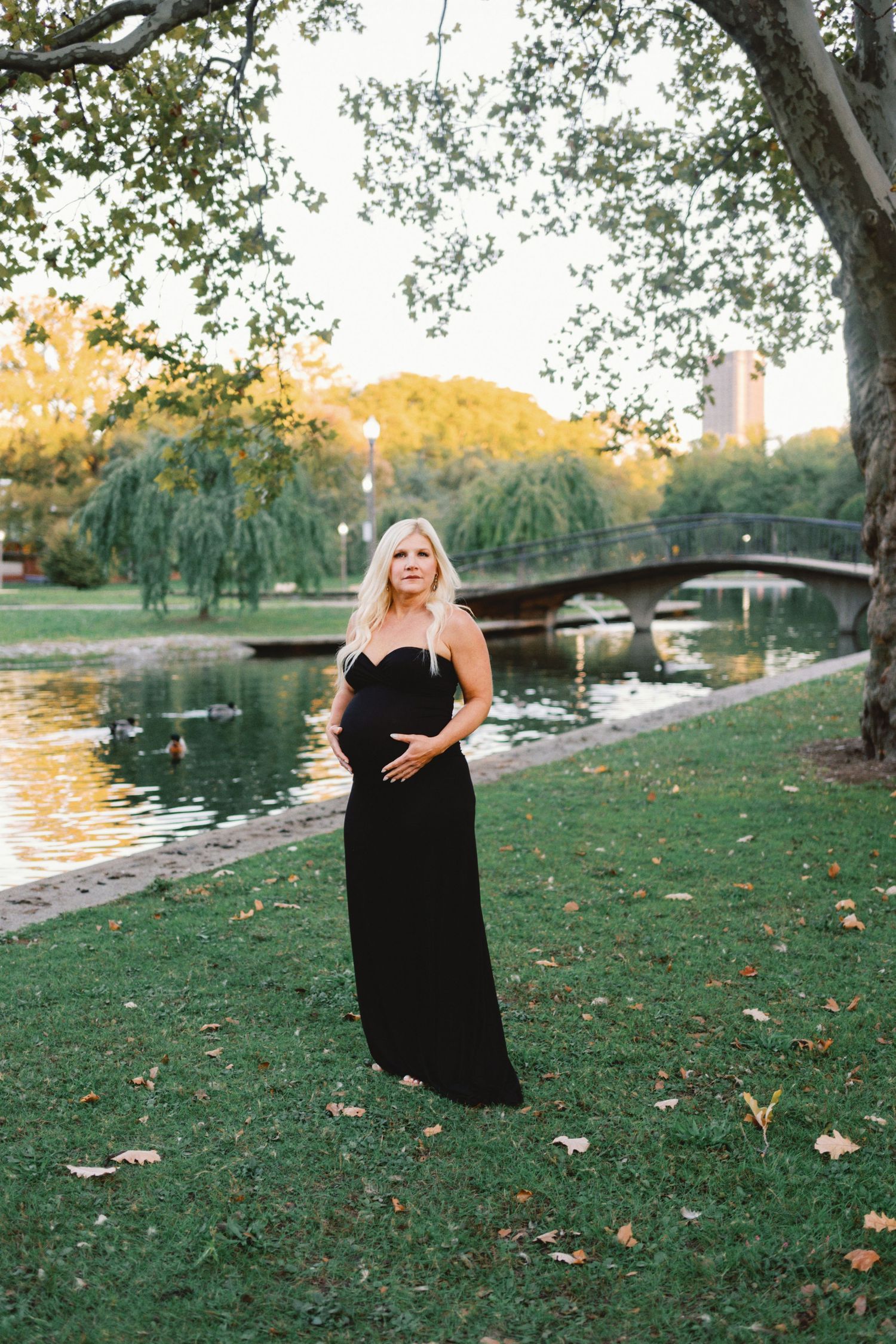 Pregnant woman in black strapless gown posing in a lush park by a pond with a bridge and willow trees.