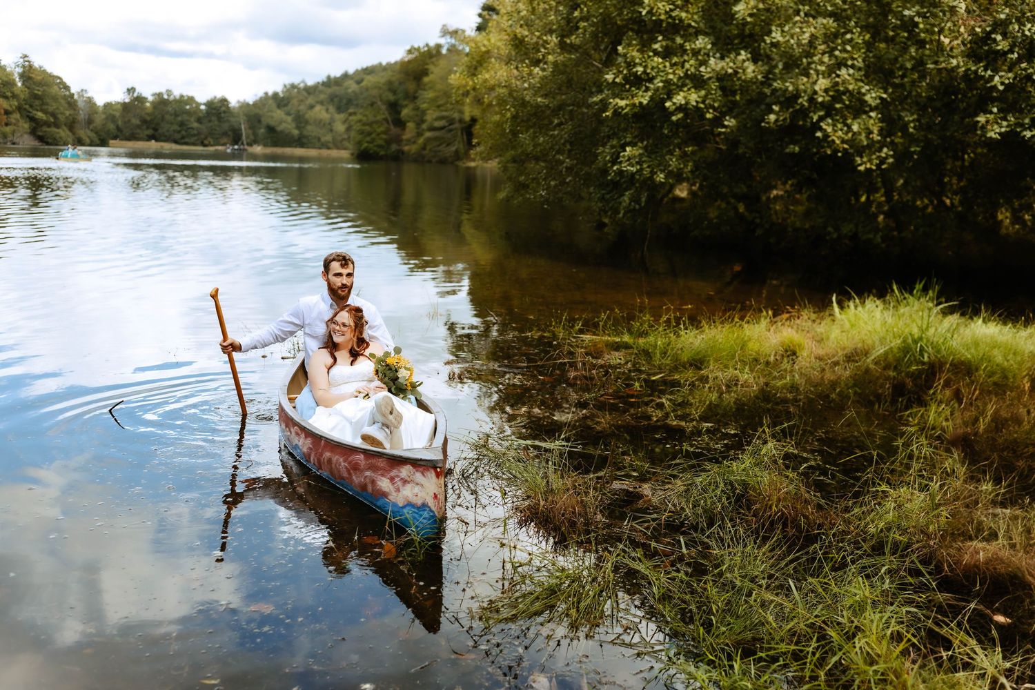 Bride and groom in a decorated canoe on a calm lake surrounded by lush green trees on their wedding day.