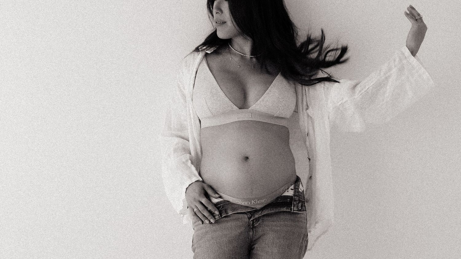 Pregnant woman in white open shirt and bralette showing baby bump in black and white maternity photo.