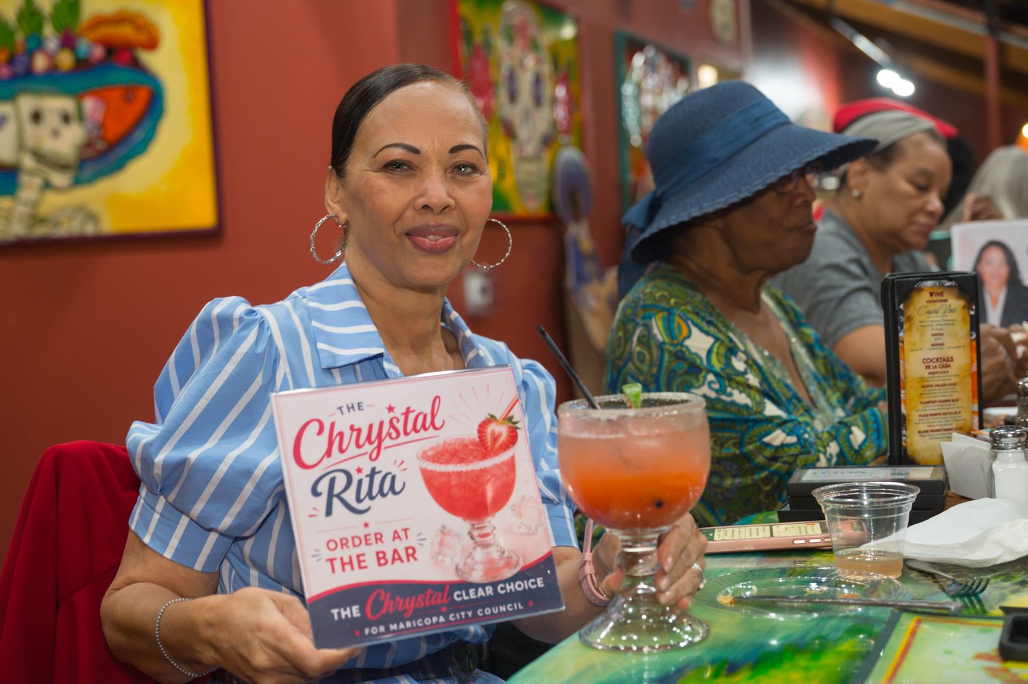Woman holding a cocktail book at a bar with a pink drink, smiling at the camera in a colorful restaurant.