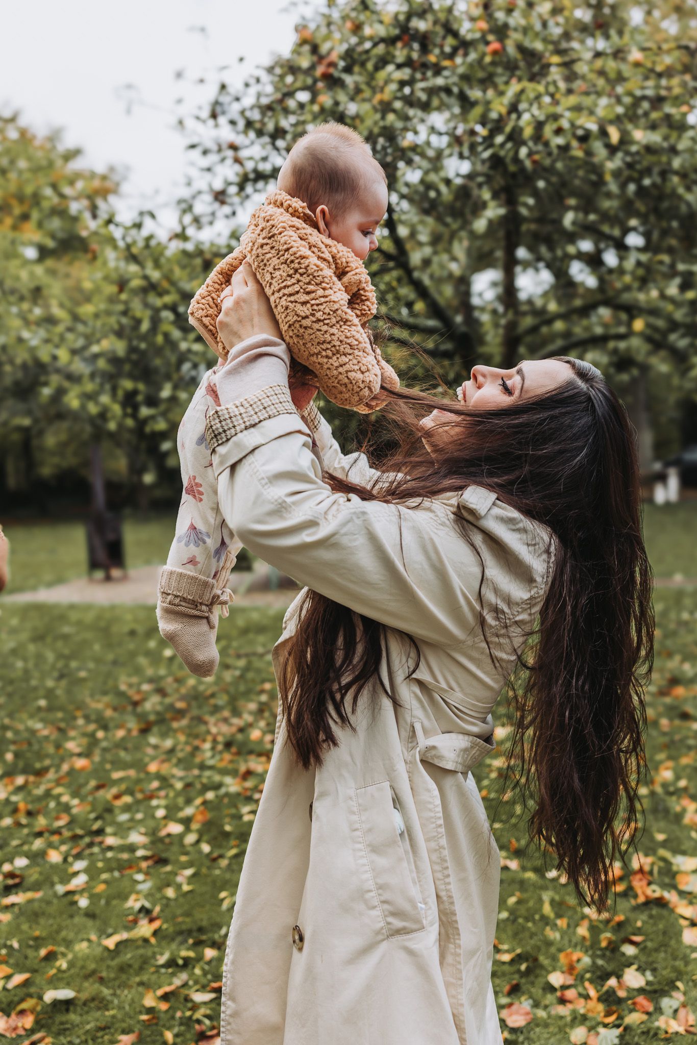 A joyful mother lifts her baby in a teddy coat outdoors in an autumn park, smiling warmly.