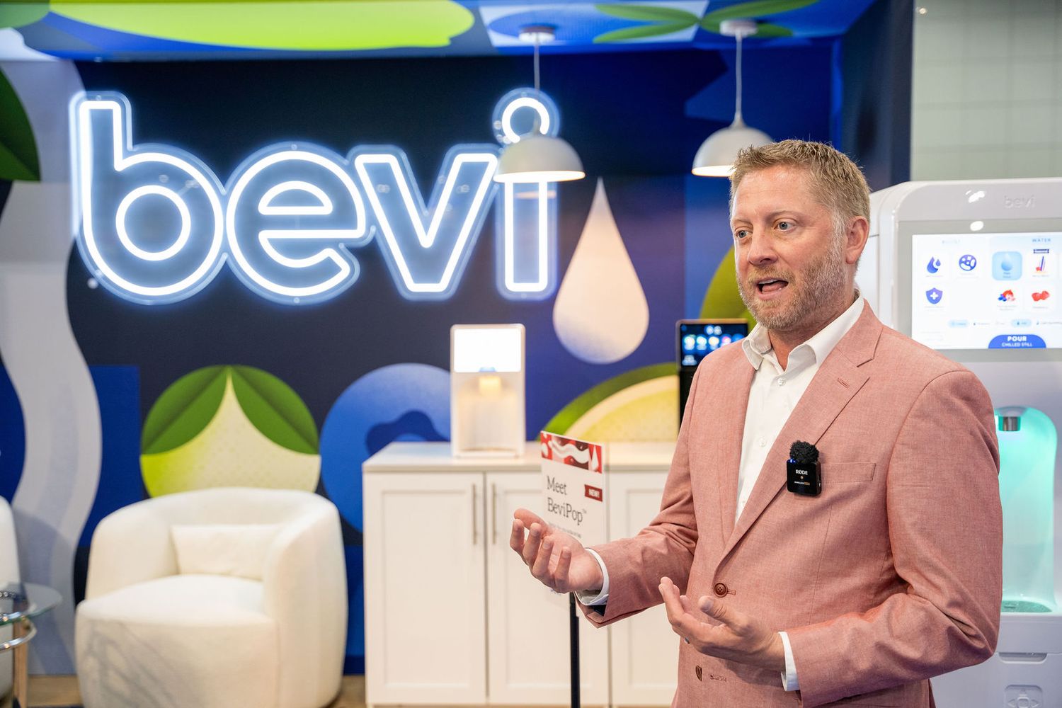 Man in pink blazer presenting Bevi smart water dispenser at a branded trade show booth display.