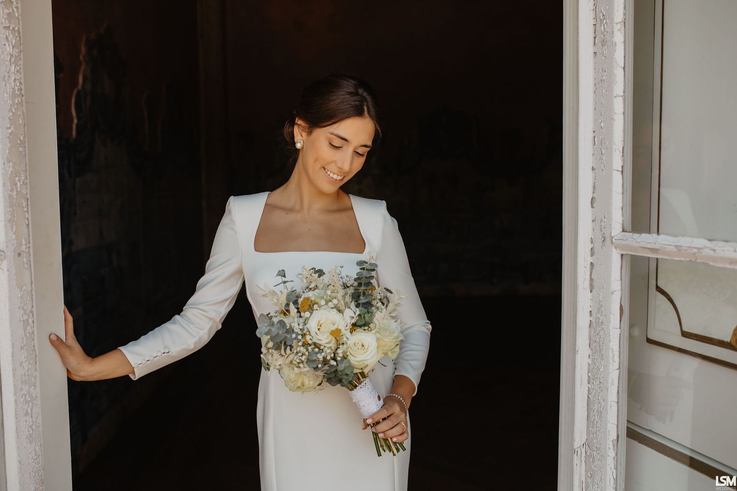 Bride in white long-sleeve gown holding bouquet of white roses and greenery, standing in elegant doorway.