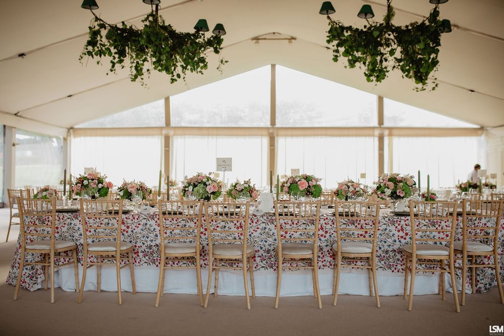 Elegant wedding reception tent with long tables, floral centerpieces, gold chairs, and lush greenery hanging from the ceiling.