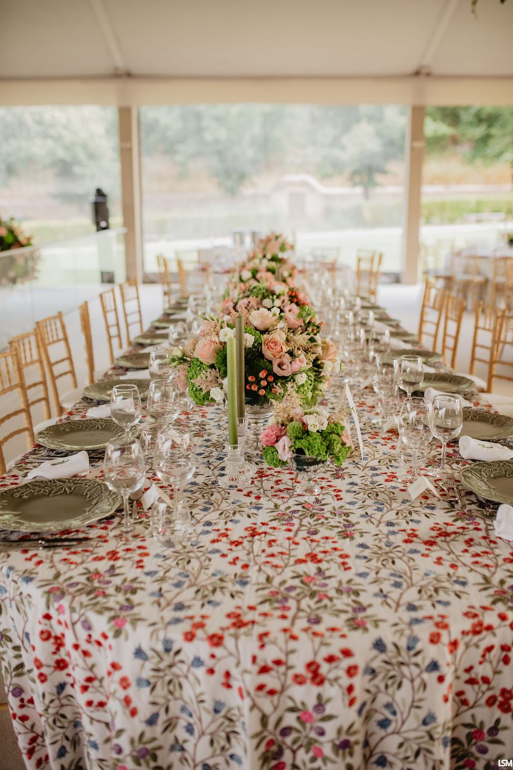 Elegant wedding reception table with floral tablecloth, gold chairs, pink flower centerpieces, and crystal glassware under a tent.
