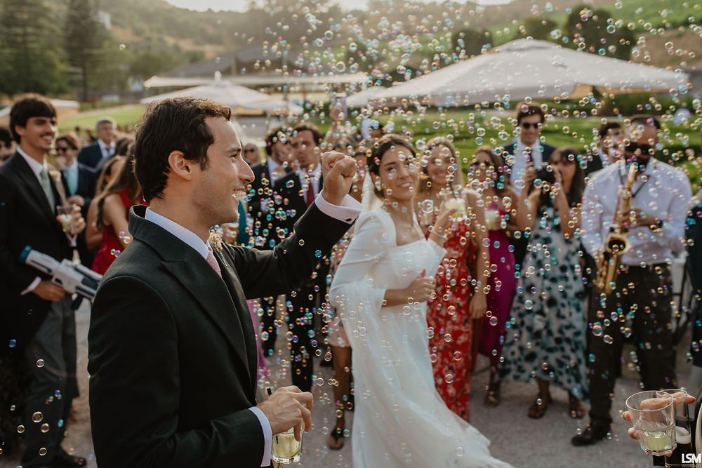 Joyful newlywed couple celebrating with bubbles surrounded by guests outdoors.