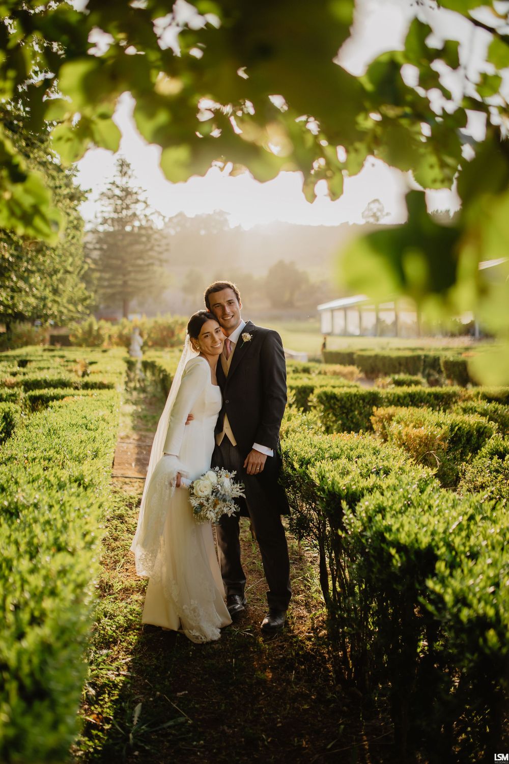 Bride and groom posing in a sunlit garden maze with lush green hedges and golden backlight.