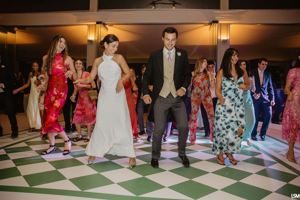Bride and groom dancing joyfully on a checkered floor surrounded by guests at a wedding reception.