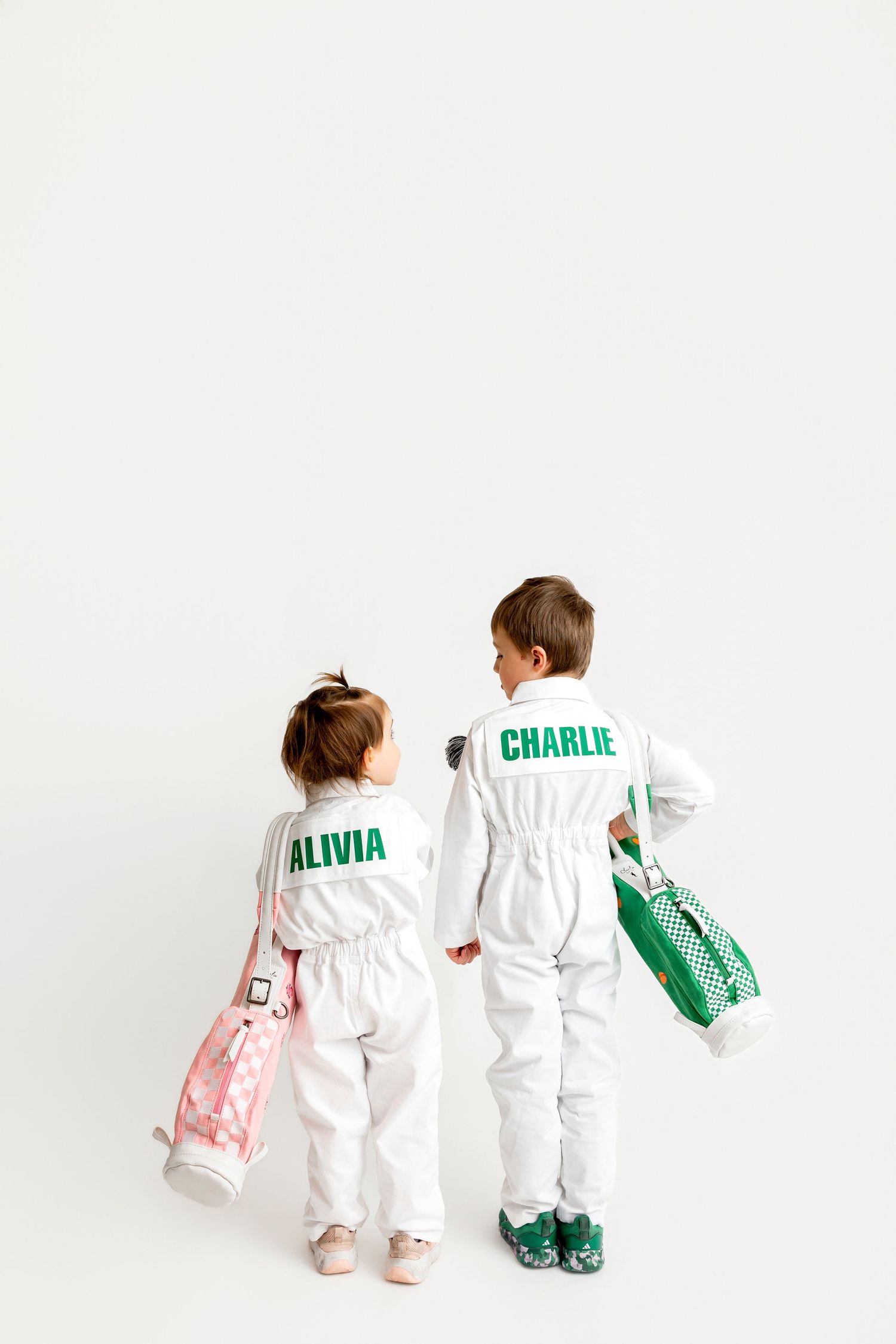 Two children named Alivia and Charlie wearing personalized white jumpsuits, holding colorful bags, viewed from behind.