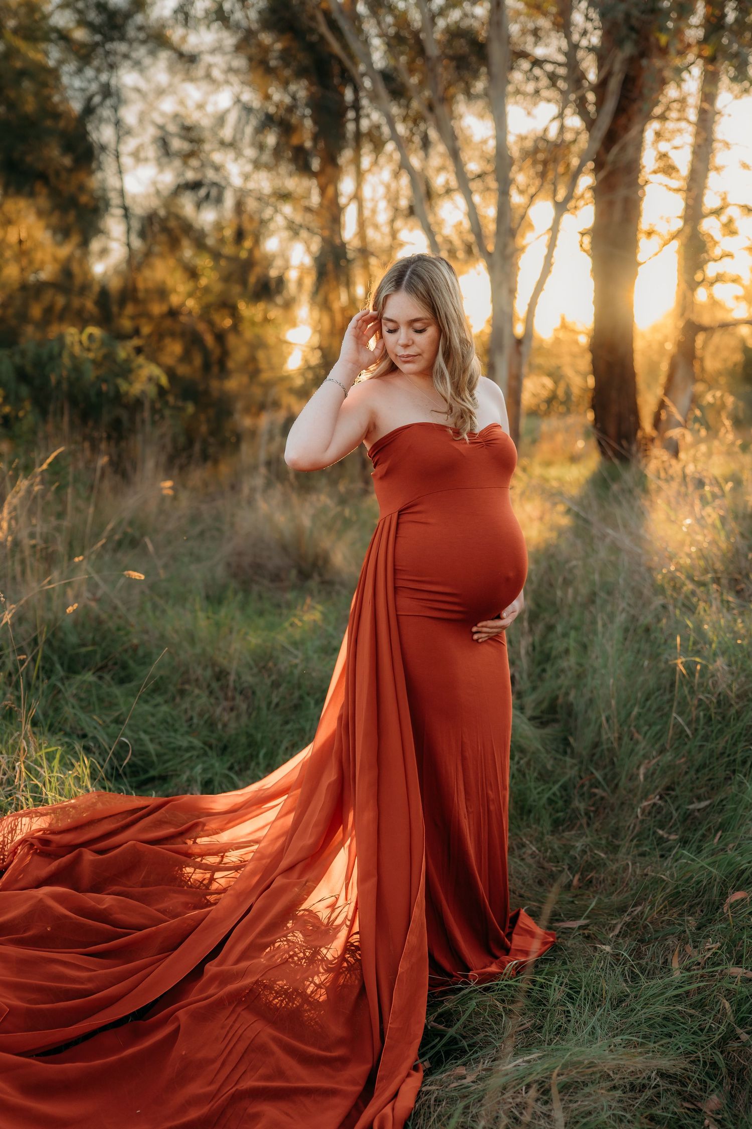 Pregnant woman in elegant rust-orange off-shoulder gown with long train poses in golden sunset forest setting.