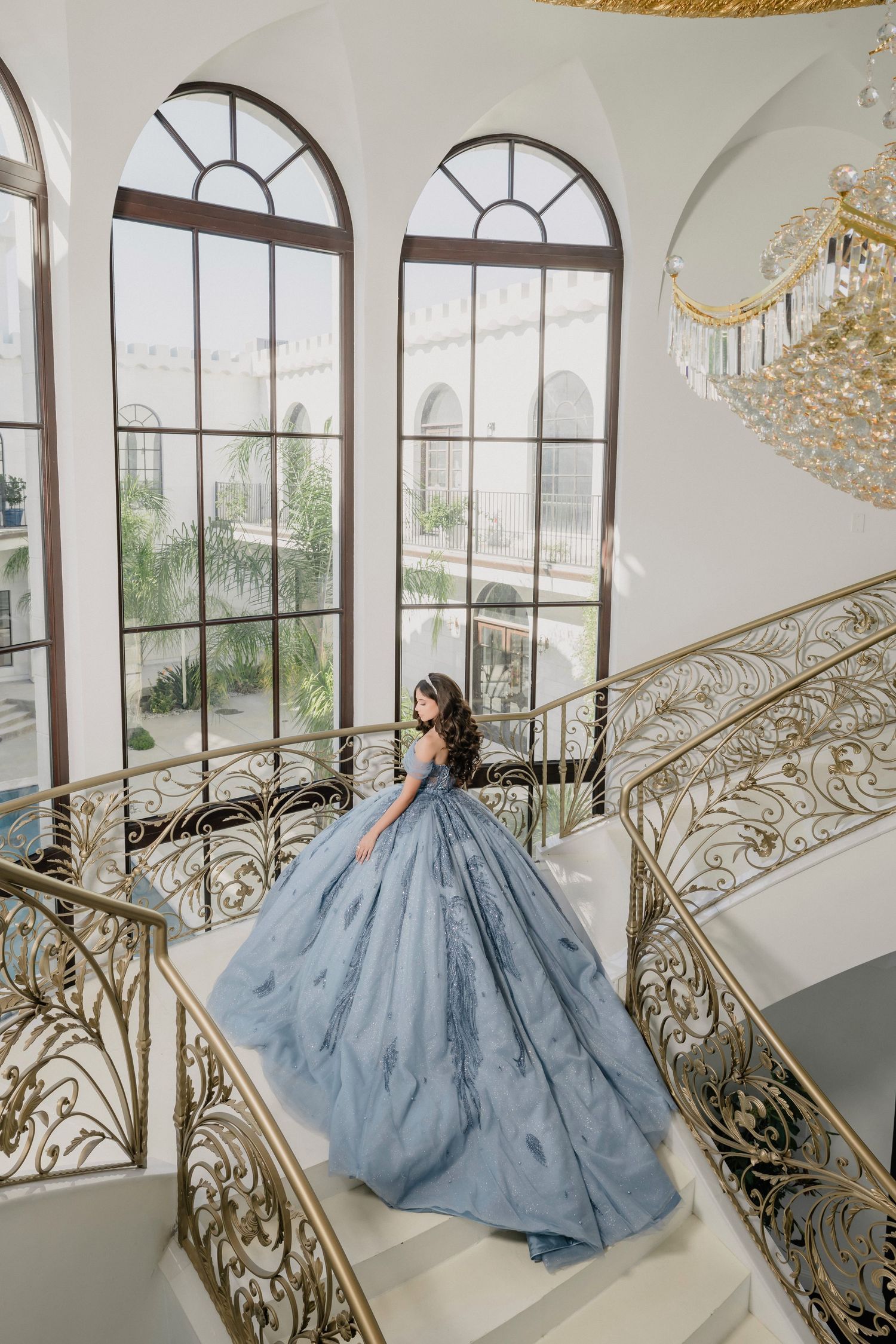 Woman in blue ballgown on ornate gold staircase in elegant mansion with arched windows and chandelier.
