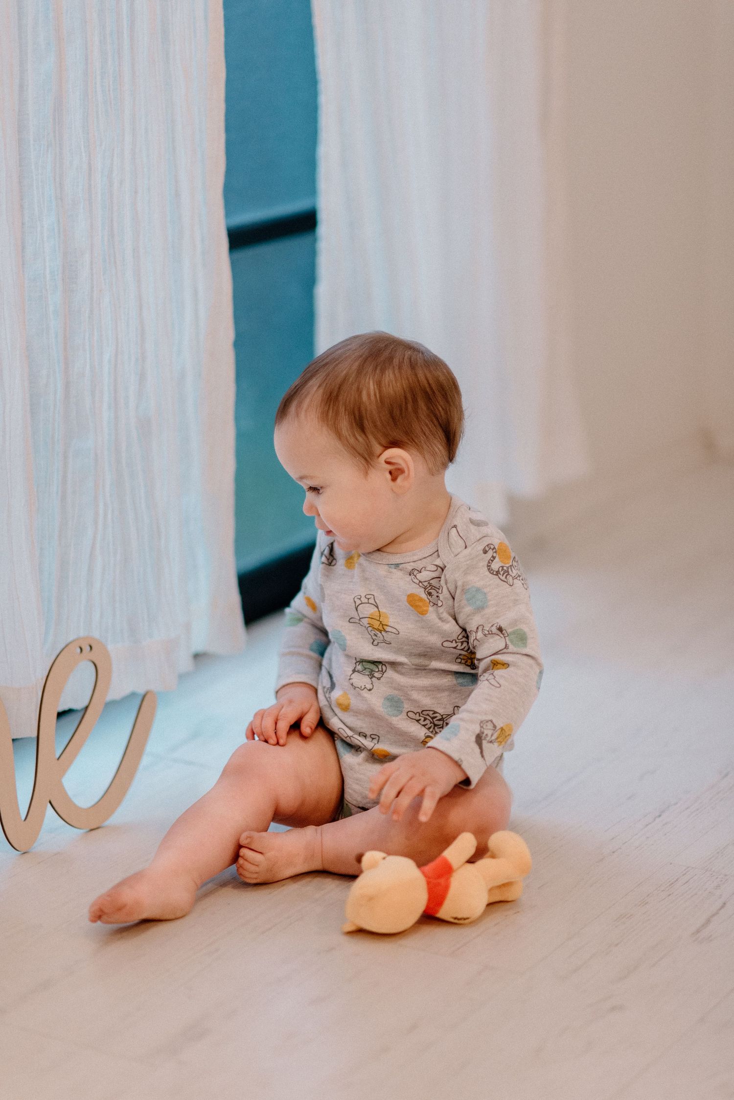 Baby in animal print onesie sitting on floor playing with stuffed giraffe toy near white curtains.