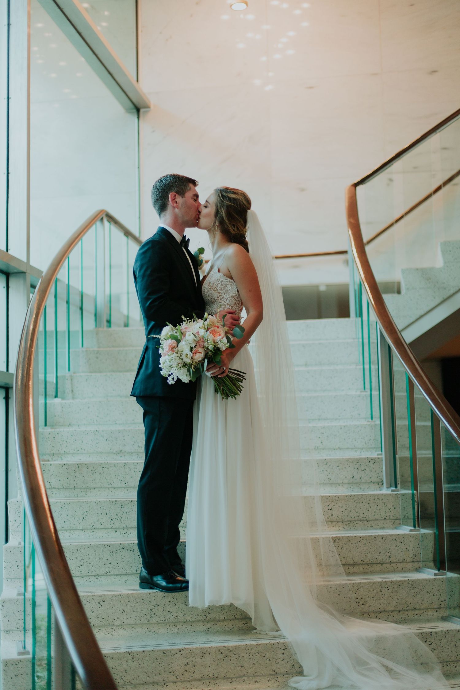 Bride and groom share a kiss on elegant staircase, bride holding pink floral bouquet with veil flowing, wedding portrait.