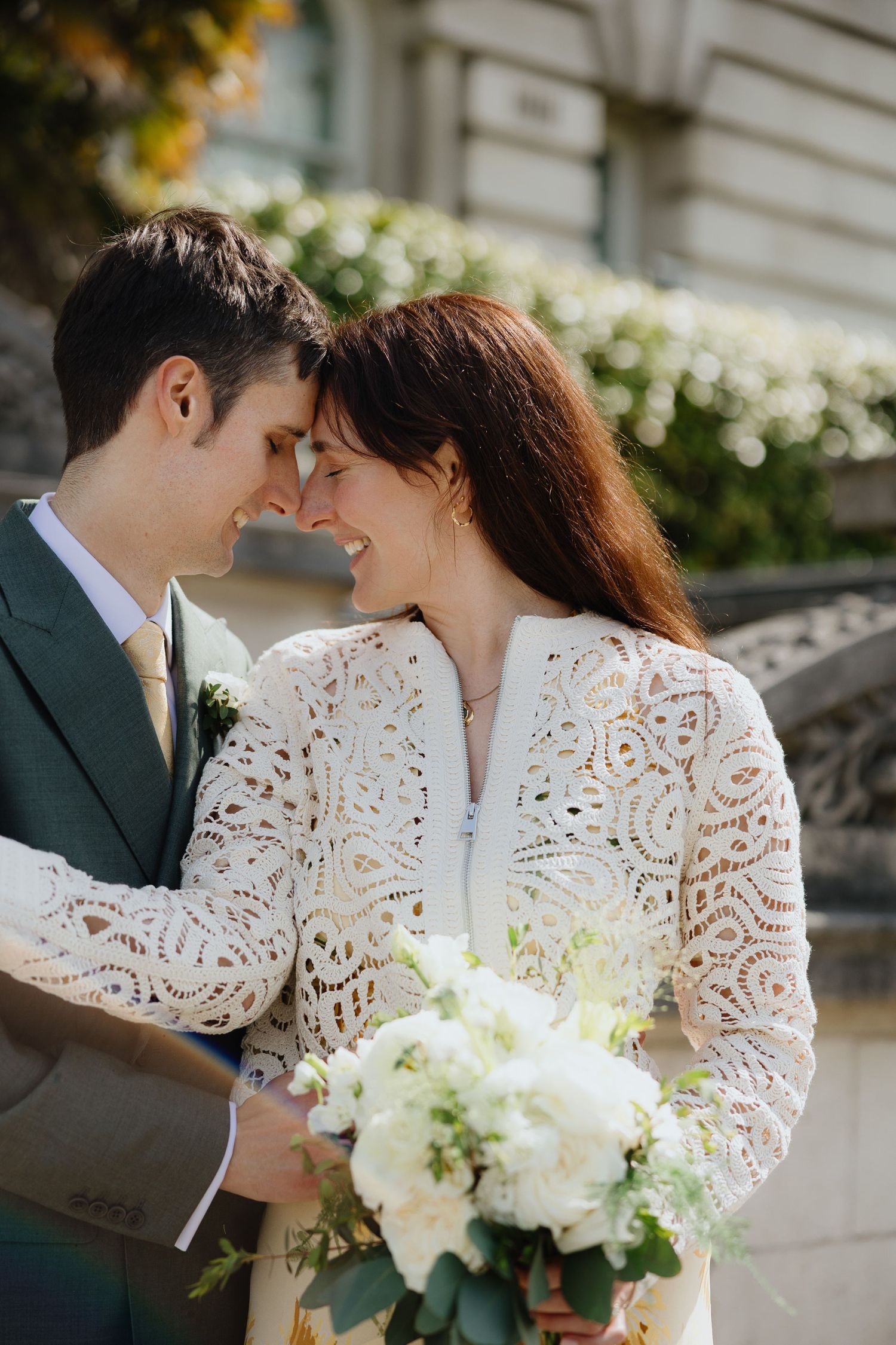 Bride and groom touching foreheads and smiling, bride holding white floral bouquet, wearing lace dress outdoors.