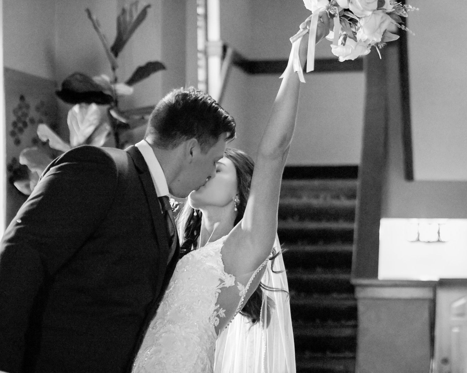 Black and white photo of a newlywed couple sharing their first kiss, bride raising bouquet in joy.