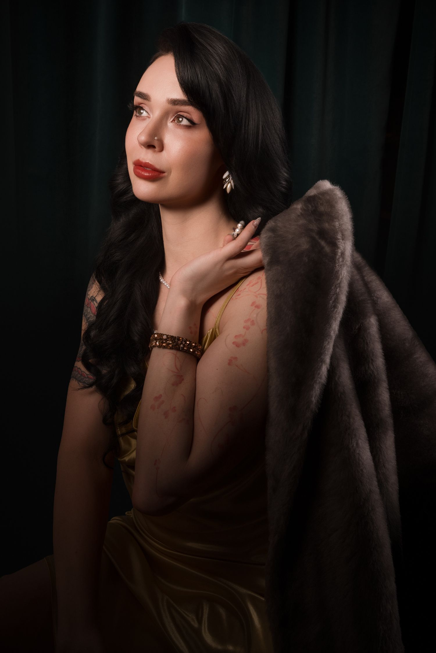 Elegant brunette woman in gold dress with fur stole, red lips, pearl earrings, posed against dark green backdrop.