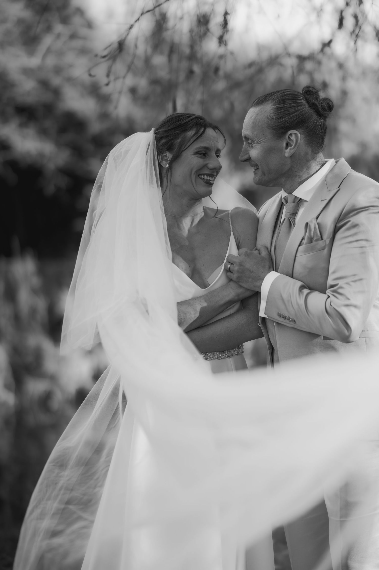 Photo en noir et blanc d'une mariée et d'un marié riants s'embrassant à l'extérieur, son voile flottant dans la brise.