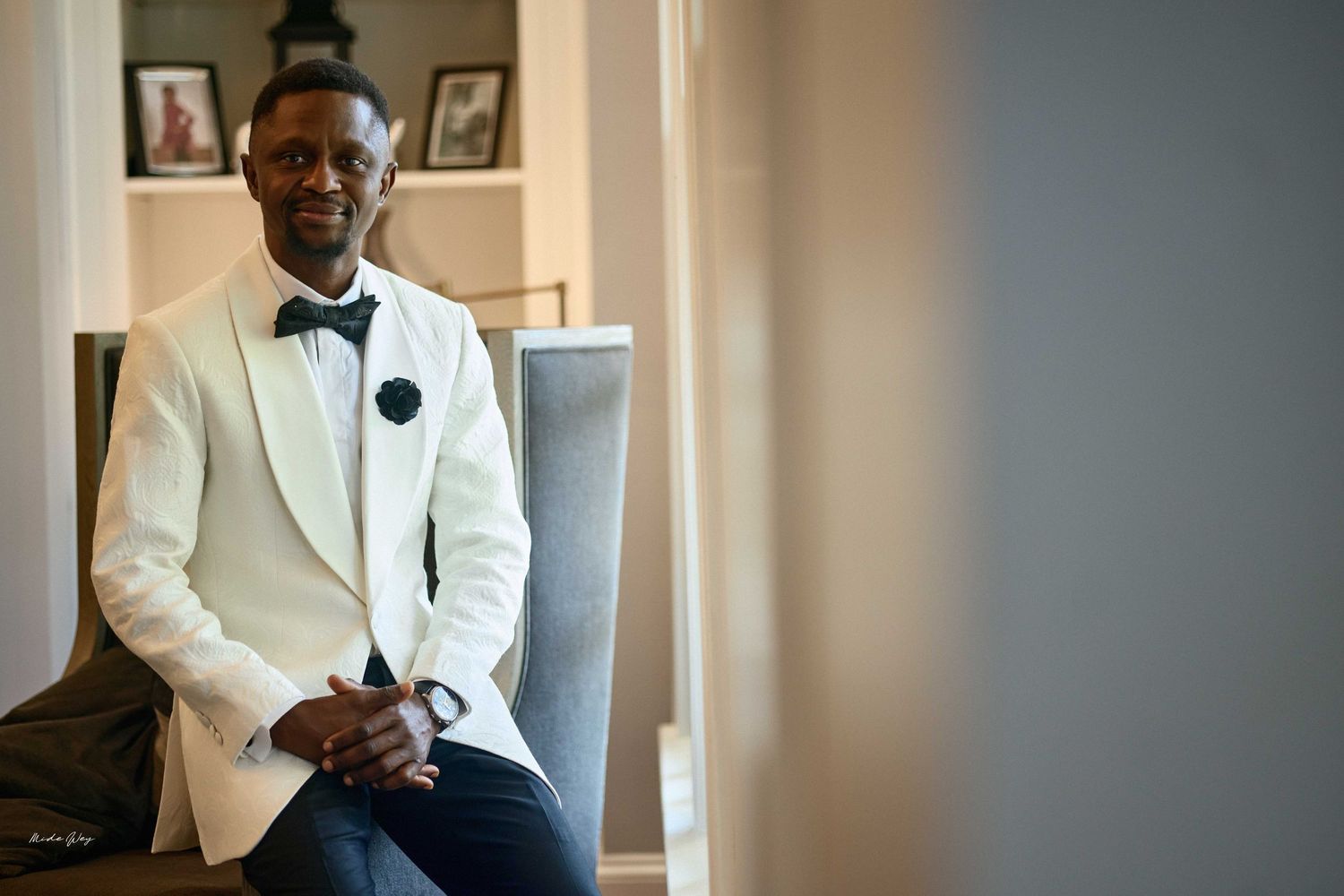 Elegant groom in white tuxedo jacket and black bow tie sitting confidently before wedding ceremony.