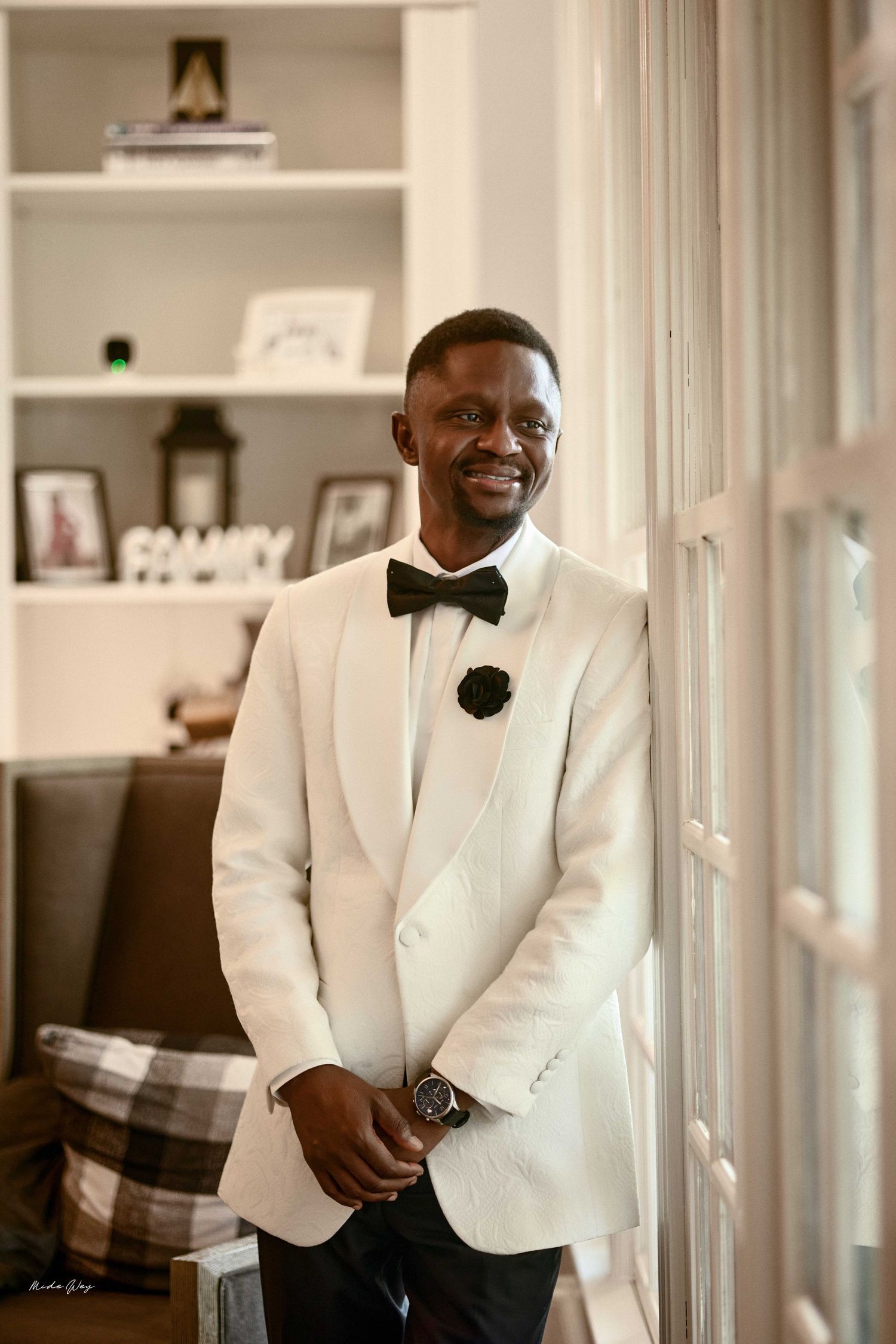 Smiling groom in white tuxedo jacket and black bow tie posing by window on wedding day.