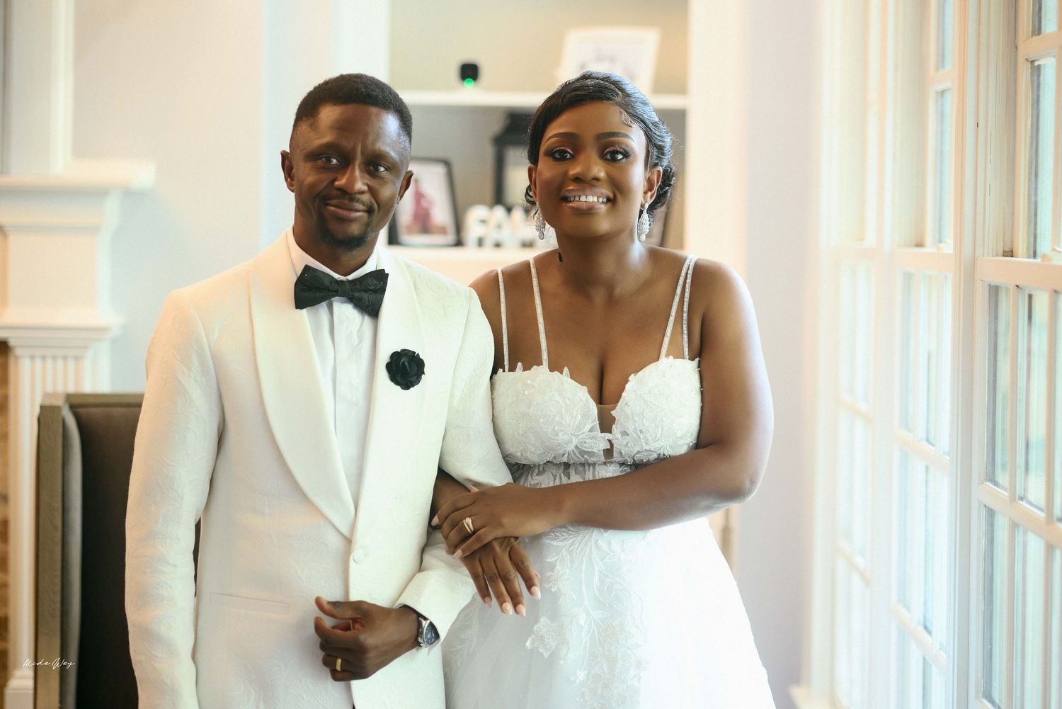Black bride and groom in white tuxedo pose elegantly together near bright windows on wedding day.
