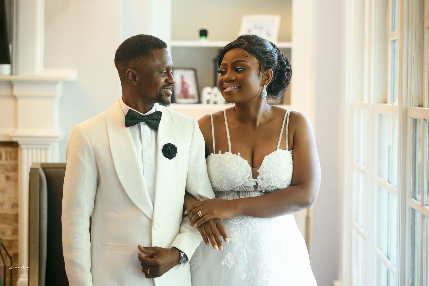 A bride and groom in elegant white wedding attire smile lovingly at each other indoors.