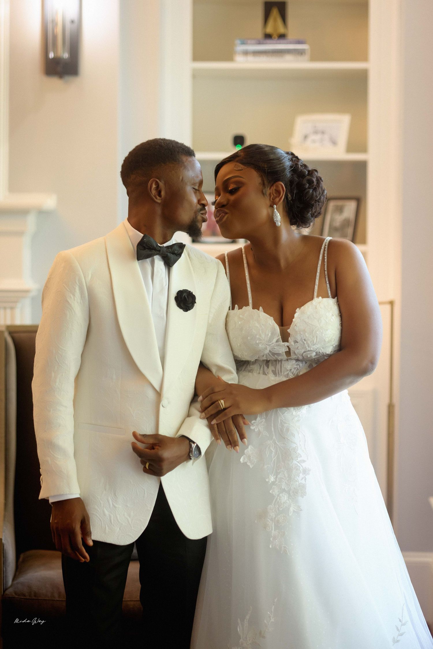 A bride and groom share a kiss in elegant wedding attire, she in a lace gown and he in a white tuxedo jacket.