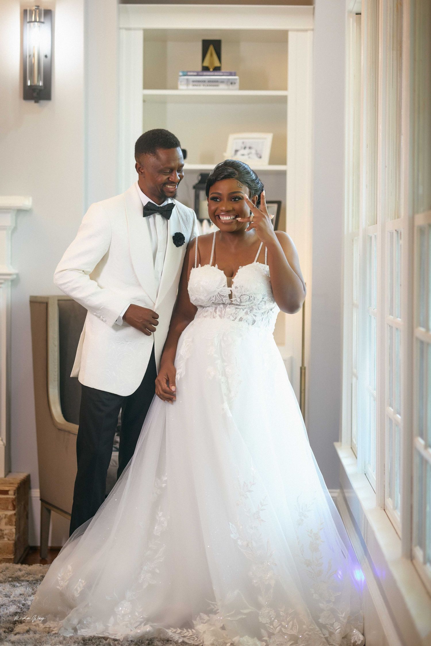 Smiling Black bride in lace wedding gown and groom in white tuxedo jacket pose together indoors.