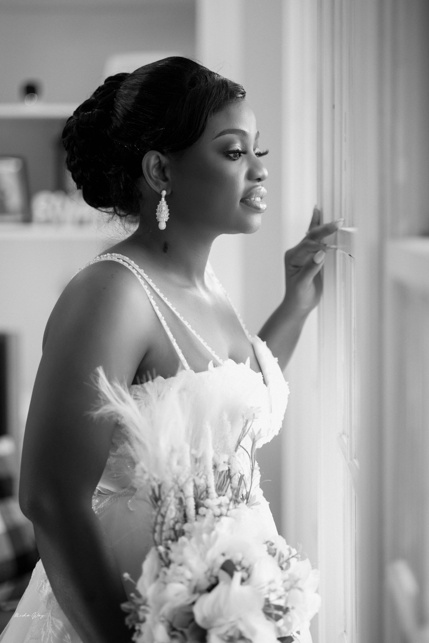 Black and white photo of a bride in feathered gown holding bouquet, gazing out window with pearl earrings.