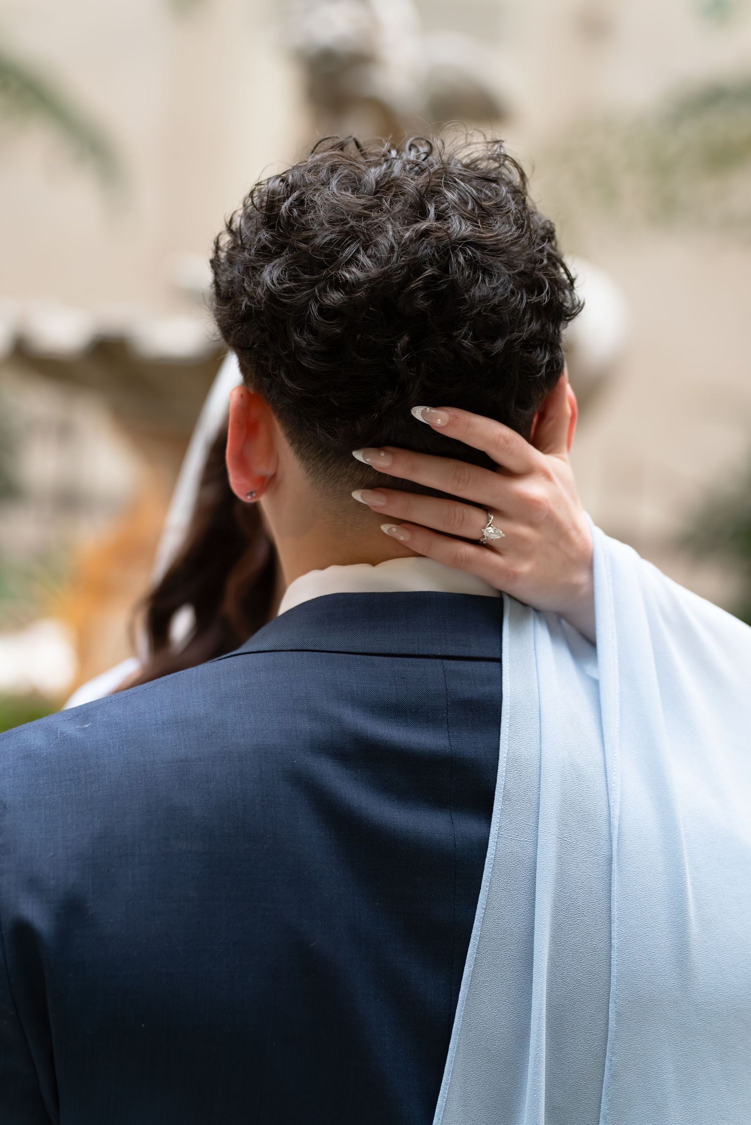 Person in light blue robe with hands covering face before a religious painting backdrop.