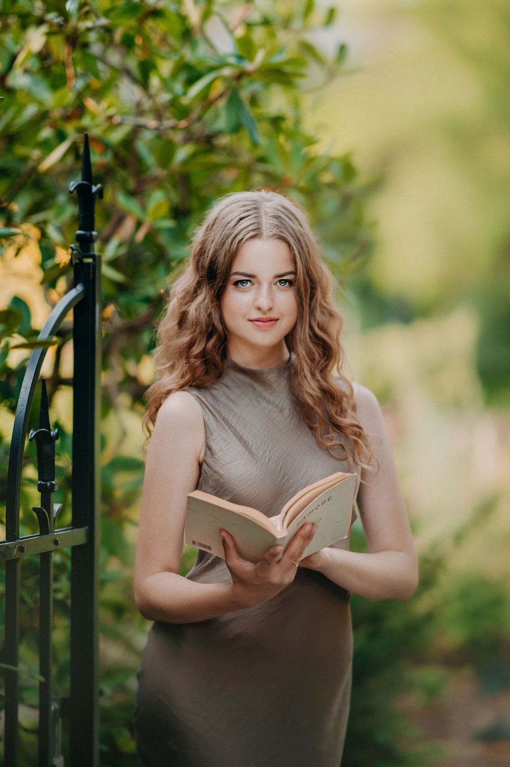 Young woman in olive dress holding open book outdoors near iron gate surrounded by lush green foliage.
