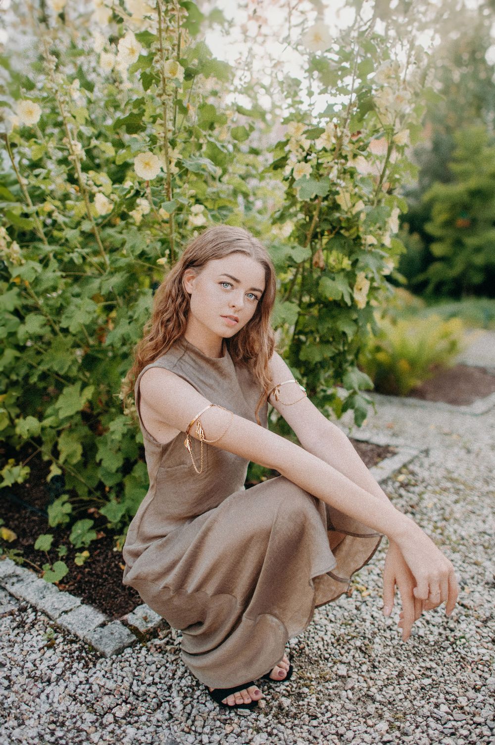 Young woman in olive linen dress crouching on gravel path surrounded by blooming hollyhock plants in garden.