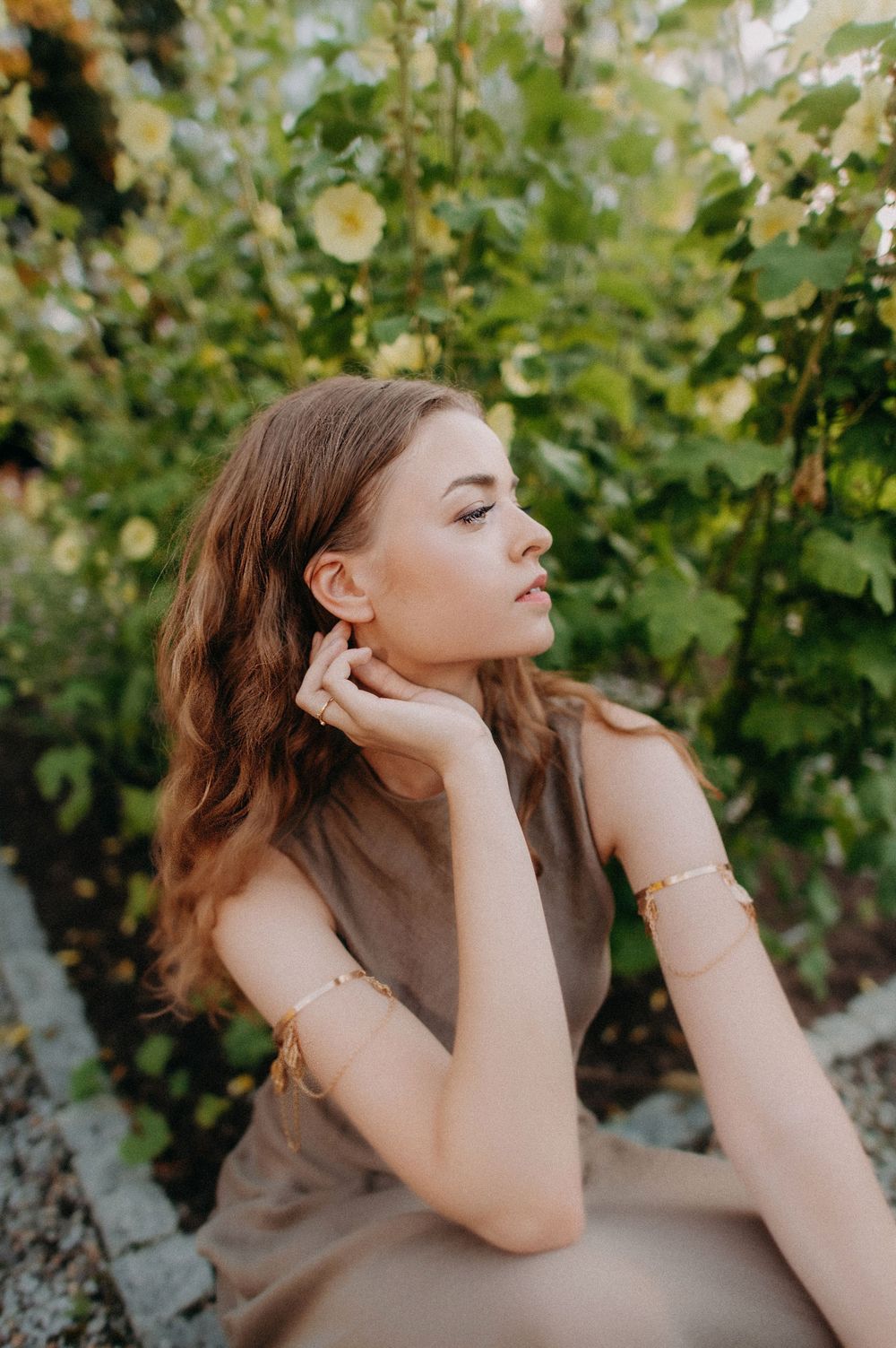 Young woman in tan dress with gold bracelets sits in a lush garden with yellow flowers, gazing thoughtfully.