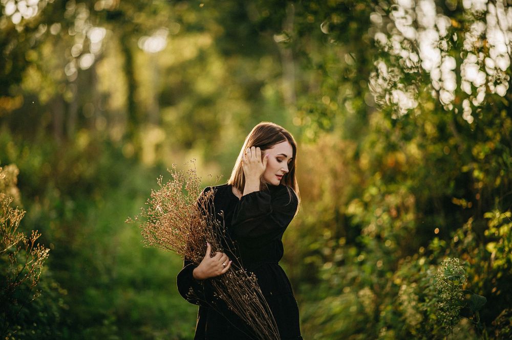 Woman in black dress holding dried wildflowers in a lush green forest path during golden hour.